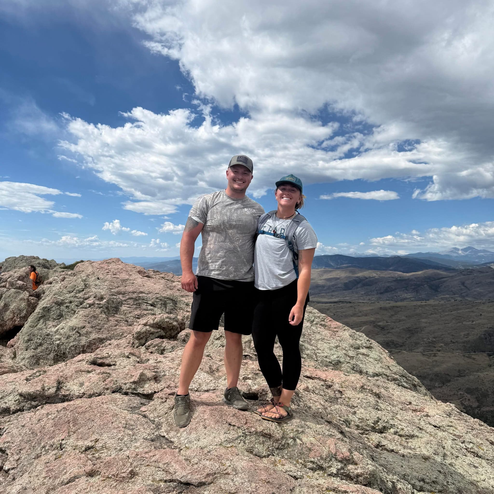 Brittany Lynn Armstrong standing on a rocky mountain summit with a wide valley and distant peaks behind her.
